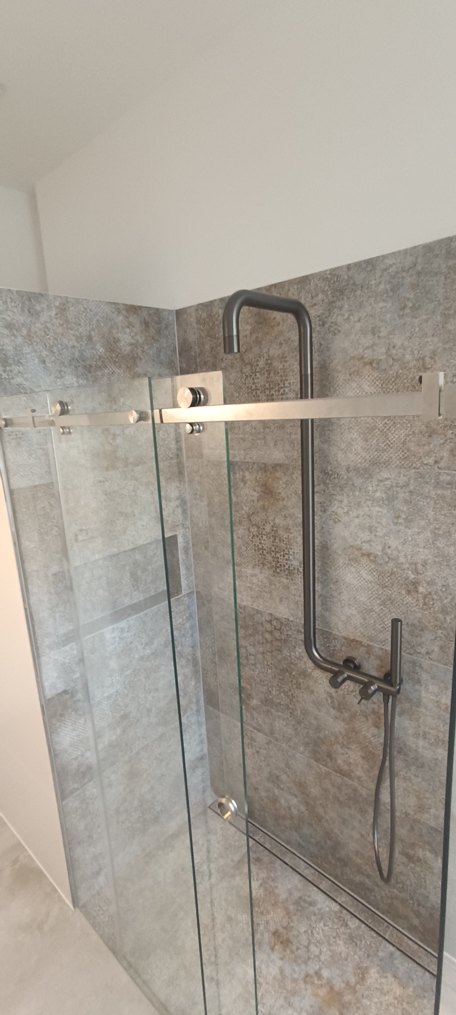 Szklana kabina prysznicowa z deszczownicą w kolorze grafitowym, zamontowana na ścianie wyłożonej płytkami imitującymi kamień w odcieniach szarości i beżu.