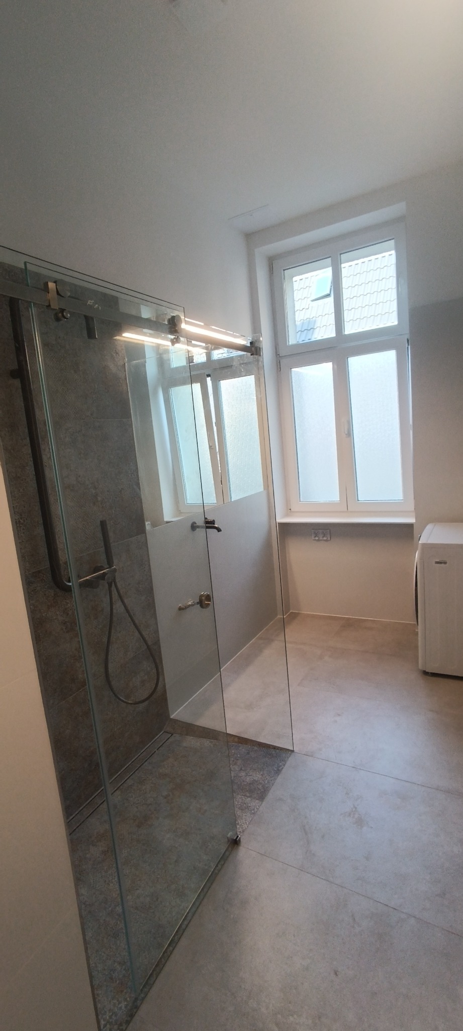 Nowoczesna kabina prysznicowa ze szklanymi drzwiami przesuwnymi, szare płytki imitujące kamień w strefie prysznica, jasne płytki podłogowe, okno z białymi ramami i widoczna pralka w tle.