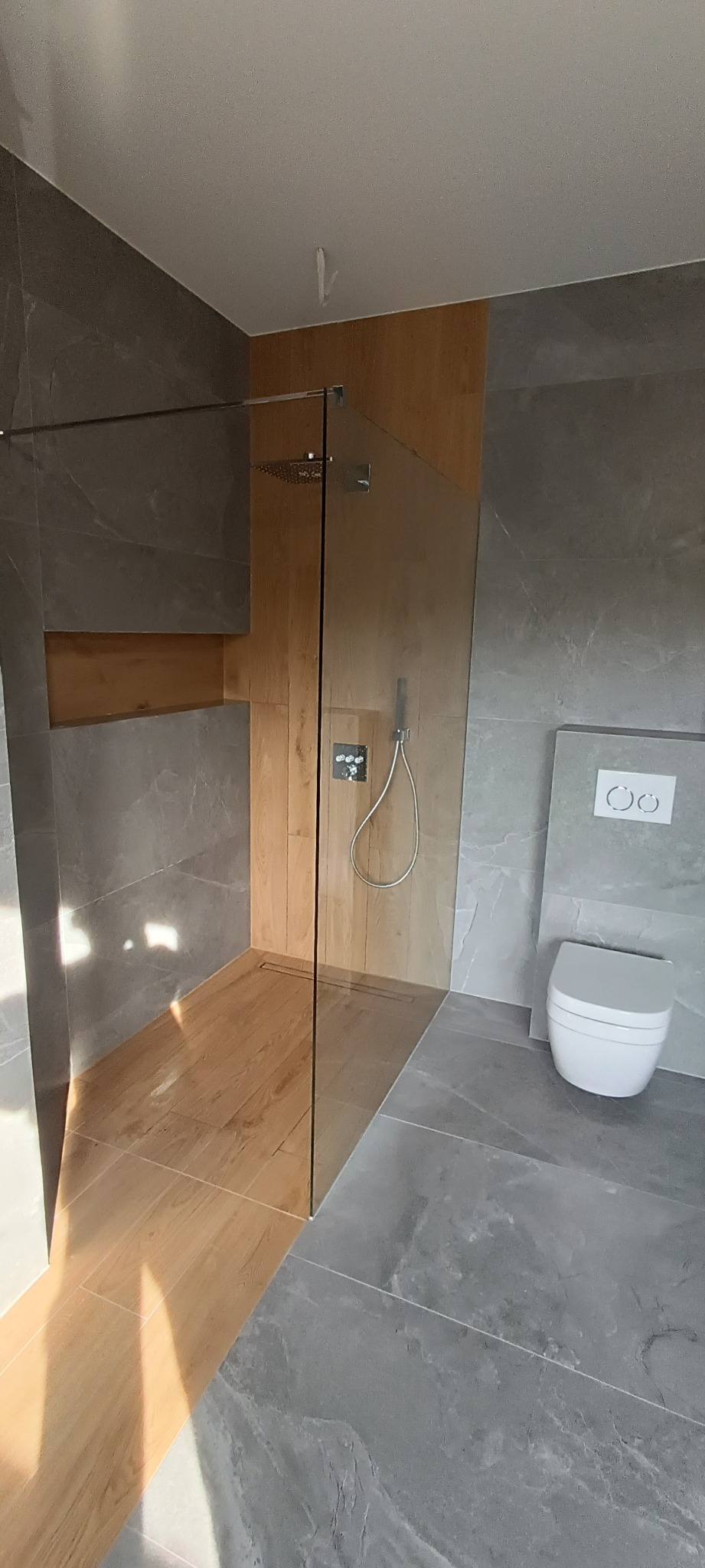 Nowoczesna łazienka z kabiną prysznicową typu walk-in, drewnopodobną podłogą w strefie prysznica i szarą marmurkową glazurą na ścianach, wisząca miska WC