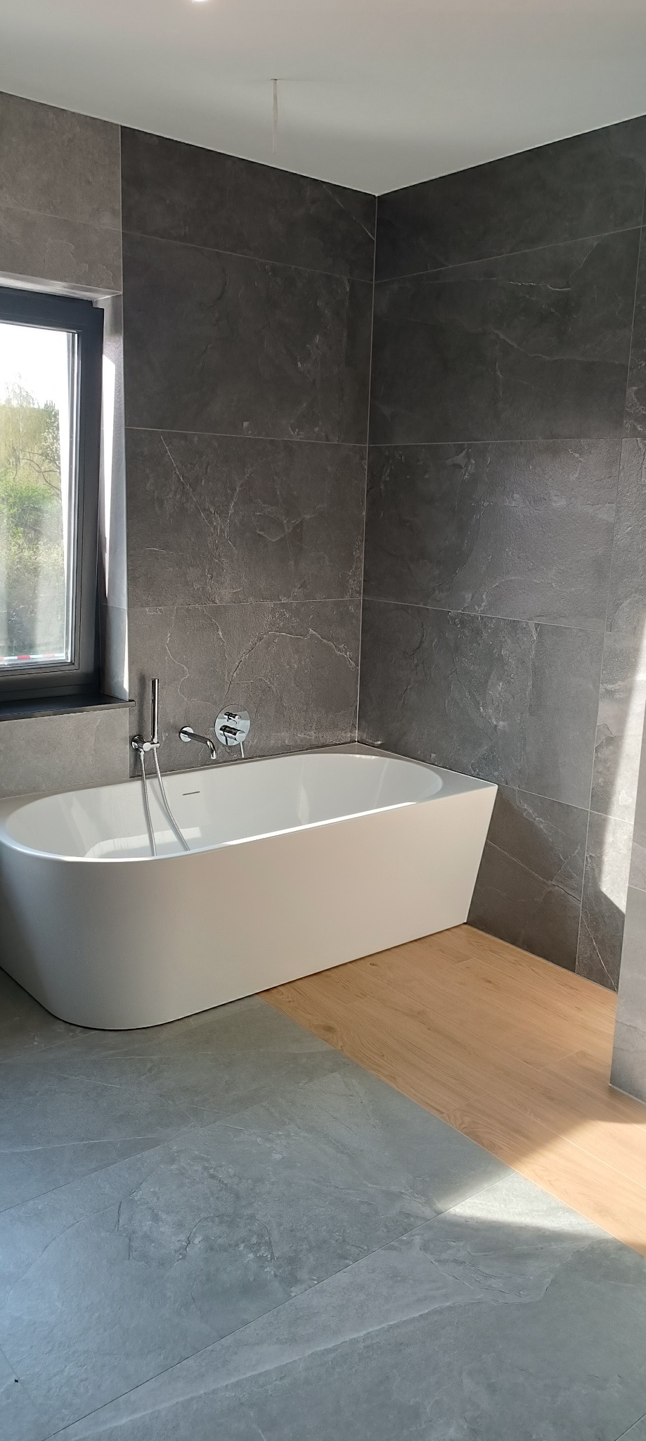 Nowoczesna łazienka z wolnostojącą białą wanną, szarymi płytkami imitującymi kamień na ścianach i podłodze, oraz fragmentem drewnianej podłogi, oświetlona naturalnym światłem z okna.
