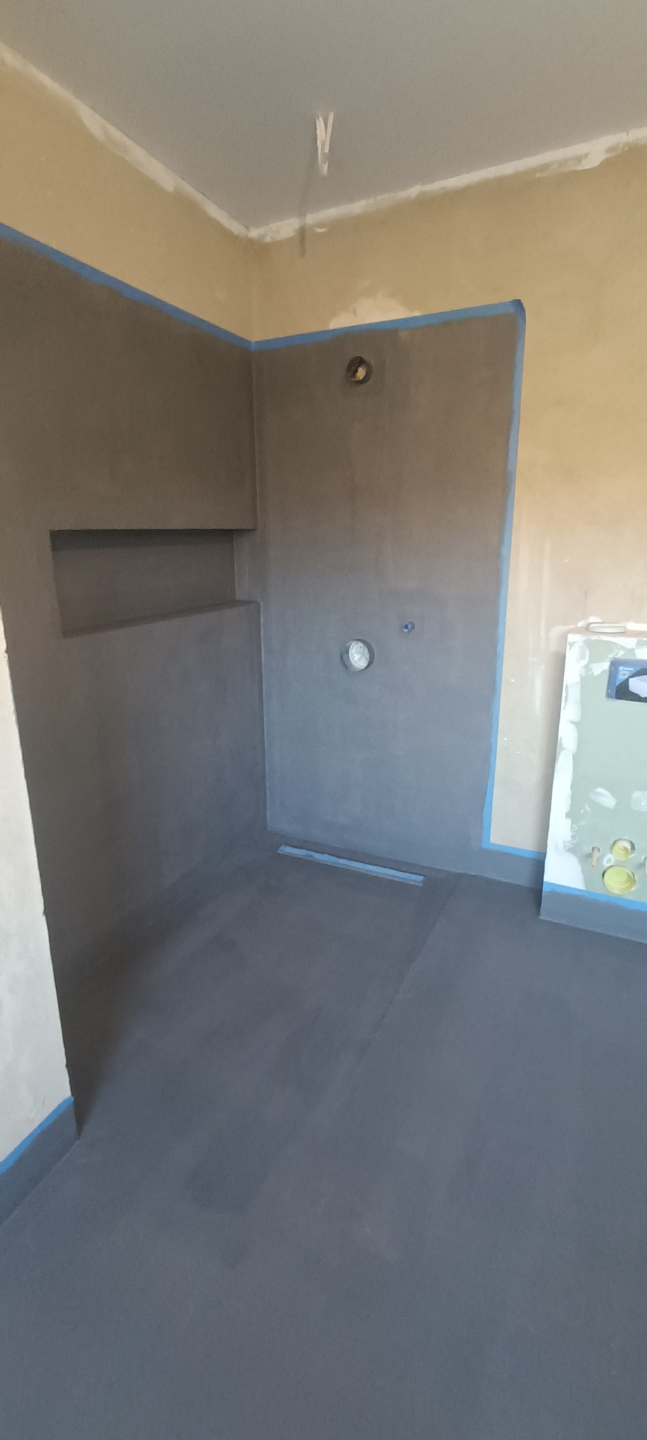 Surowa łazienka w trakcie wykańczania, ściany i podłoga pokryte szarą hydroizolacją, widoczna wnęka w ścianie, odpływ liniowy w podłodze, otwory na armaturę, zabezpieczone taśmą malarską krawędzie.