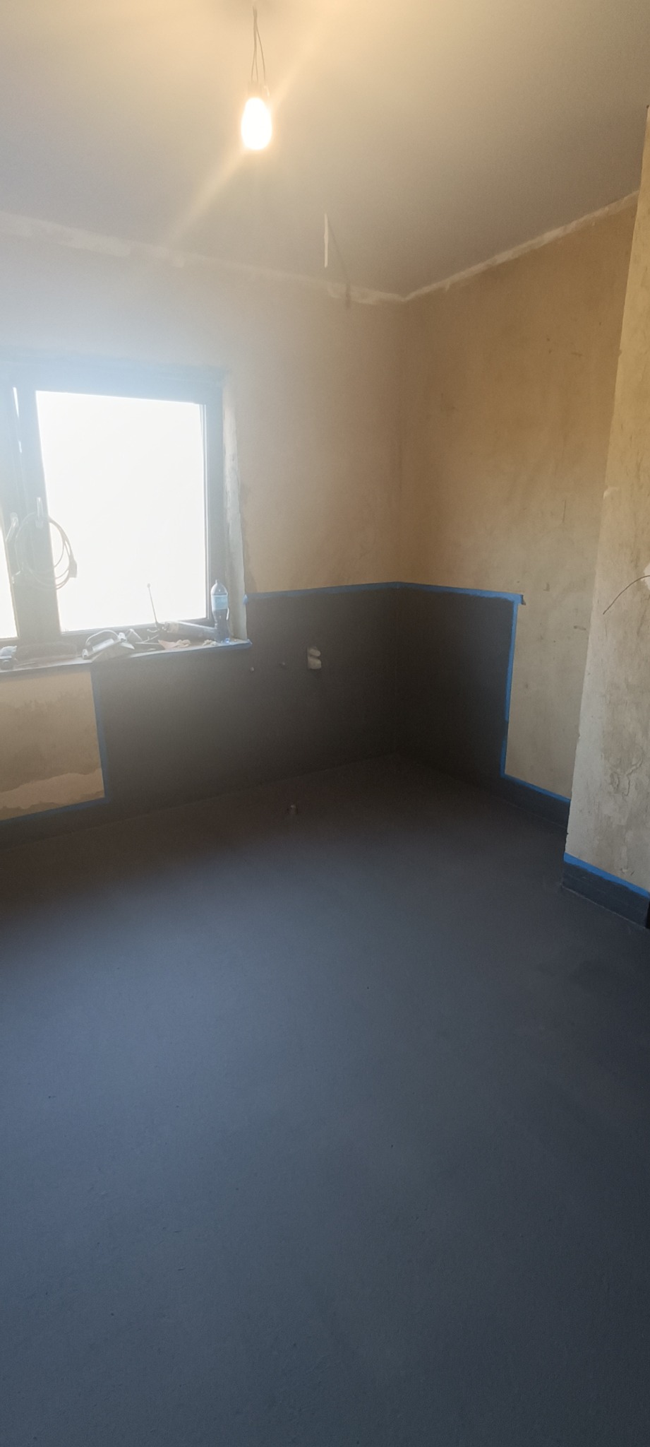 Przygotowane do dalszych prac pomieszczenie z wylaną, ciemnoszarą posadzką, zabezpieczonymi taśmą malarską ścianami i oknem z widocznymi narzędziami.