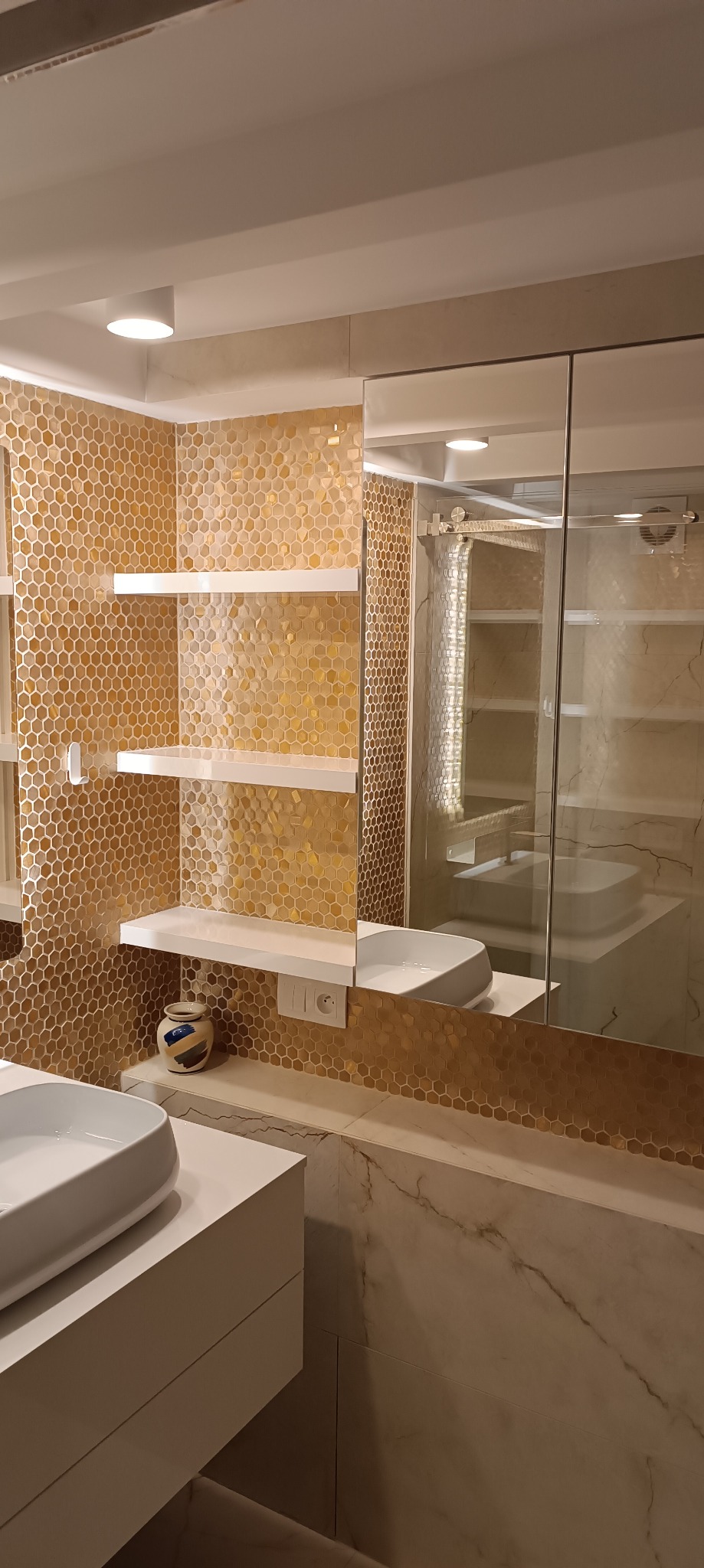 Nowoczesna łazienka z mozaiką w odcieniach złota, białymi półkami i umywalką na blacie, odbicie kabiny prysznicowej w lustrze.