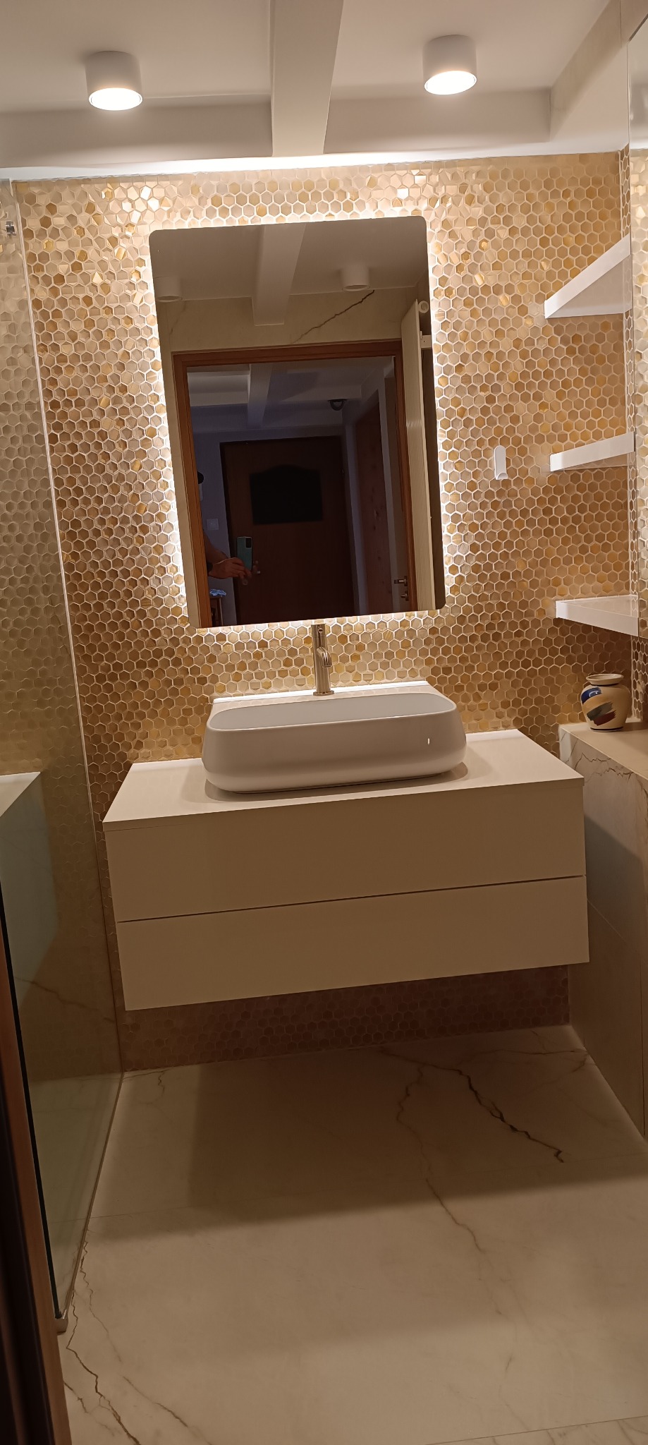 Nowoczesna łazienka z umywalką nablatową o zaokrąglonym kształcie, podświetlanym lustrem i ścianą wyłożoną mozaiką w odcieniach złota. W lustrze odbija się wnętrze mieszkania.