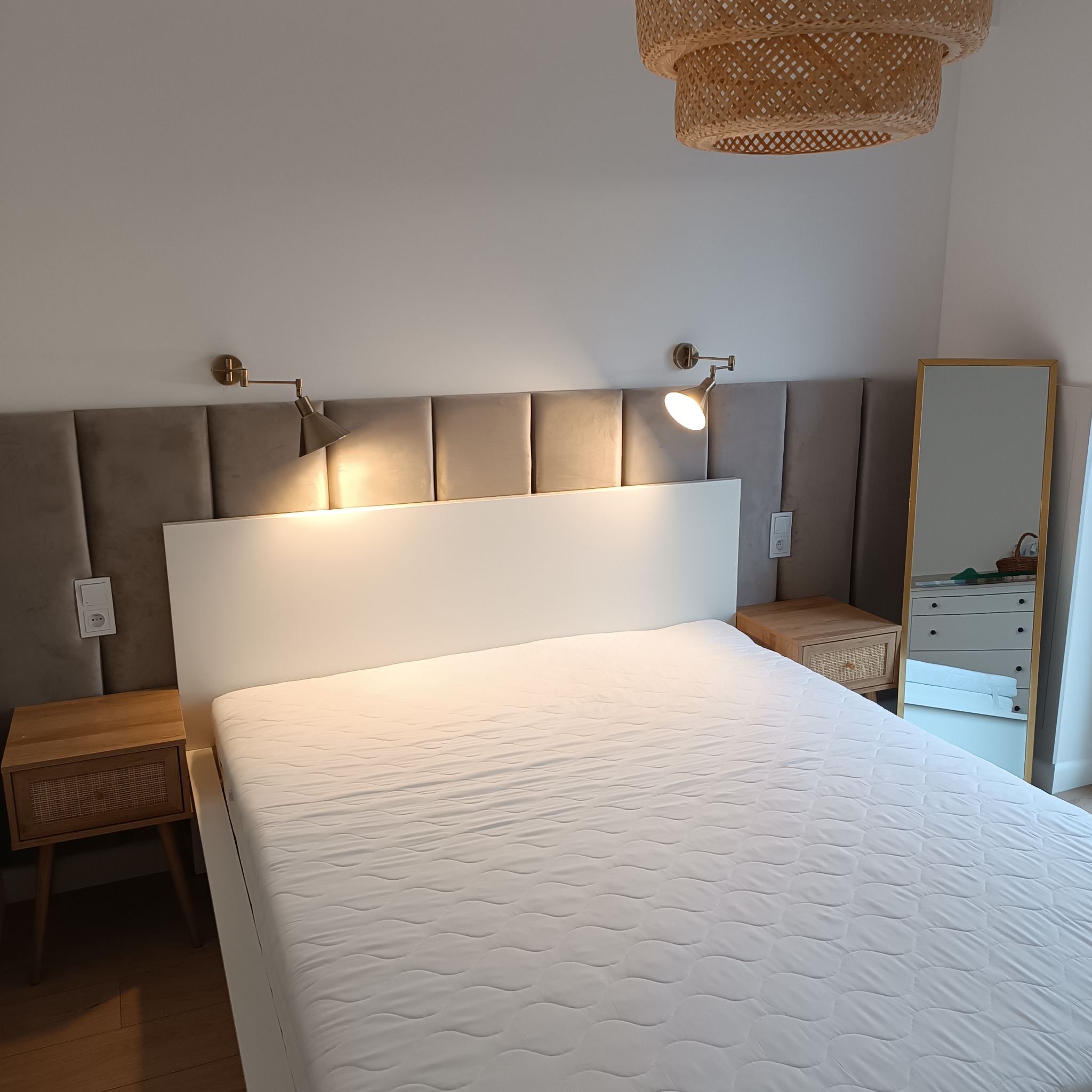 Nowoczesna sypialnia z łóżkiem z białym zagłówkiem, panelem tapicerowanym w kolorze taupe, lampkami nocnymi w stylu retro i rattanowymi szafkami nocnymi, wisząca lampa z rattanu.