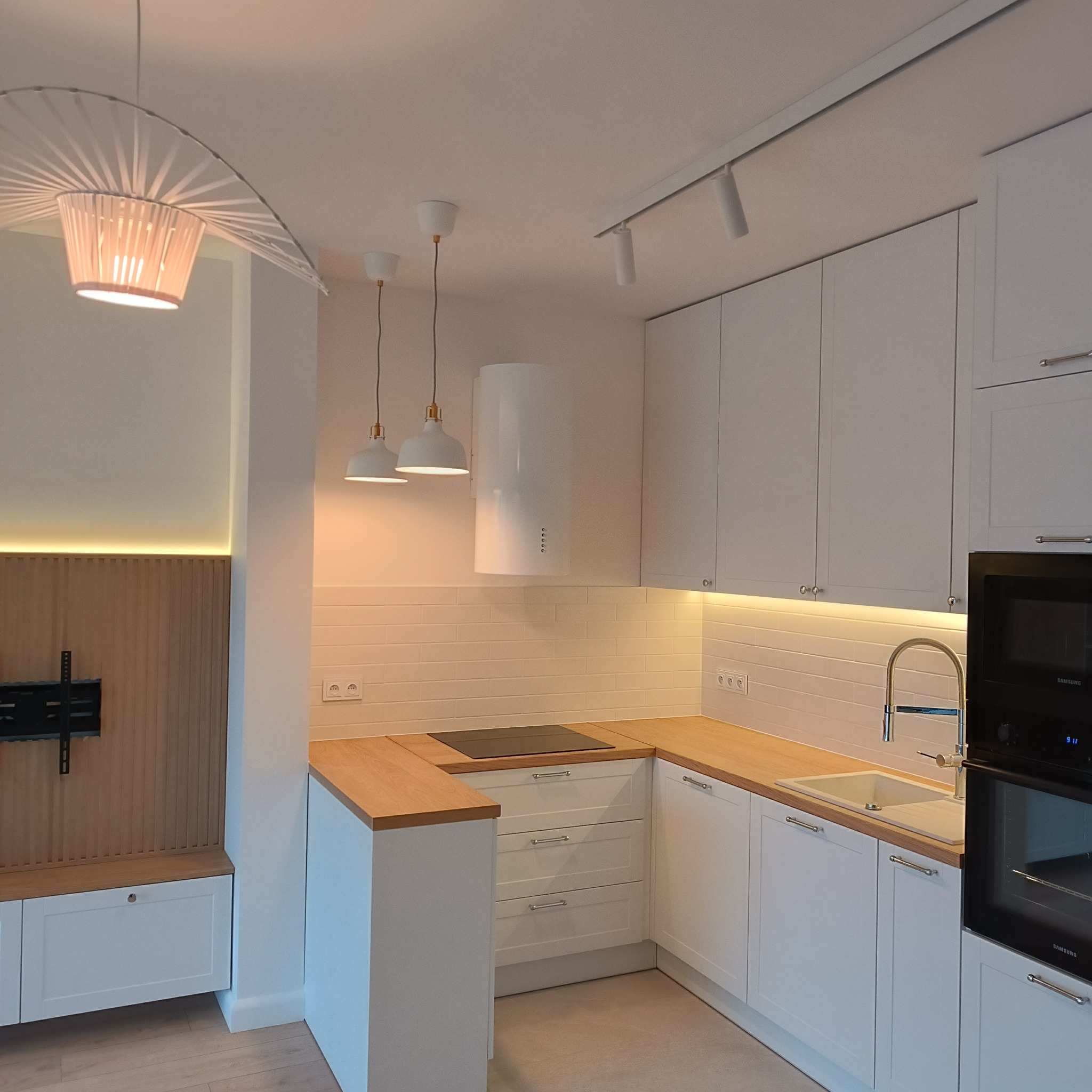Nowoczesna, jasna kuchnia z białymi szafkami, drewnianym blatem i oświetleniem LED podszafkowym, uzupełniona minimalistycznymi lampami wiszącymi i designerskim abażurem.