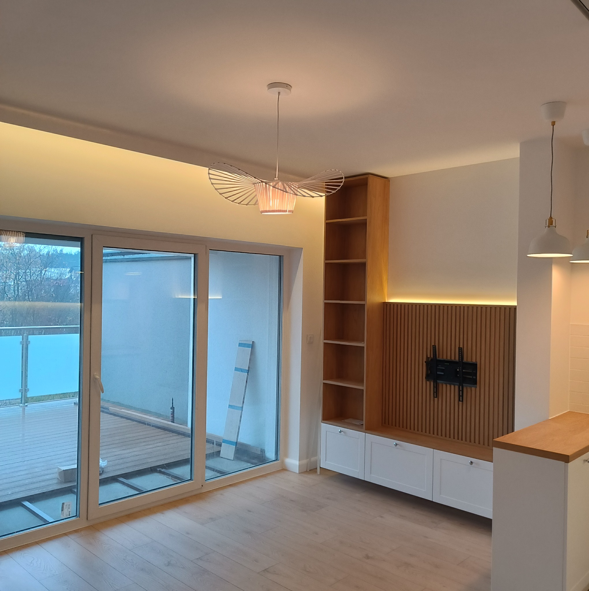 Jasne wnętrze po remoncie z drewnianą podłogą, dużymi oknami balkonowymi, zabudową meblową z drewnianymi akcentami i dekoracyjnym oświetleniem.