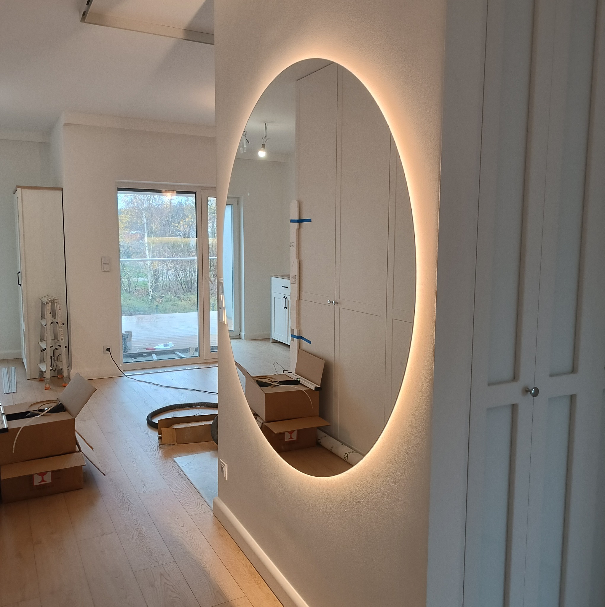 Okrągłe lustro z podświetleniem LED na ścianie w trakcie montażu, widoczne kartony i narzędzia, wnętrze w trakcie prac wykończeniowych.