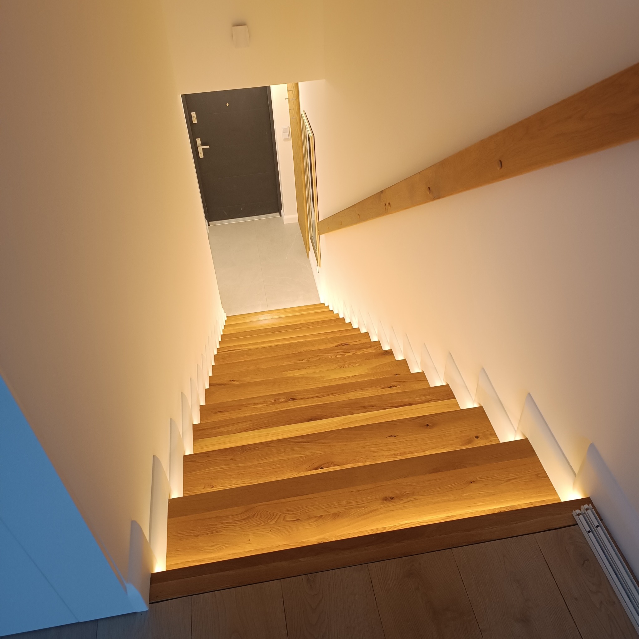 Drewniane schody z podświetleniem LED wzdłuż ściany, widok z góry, minimalistyczny design.