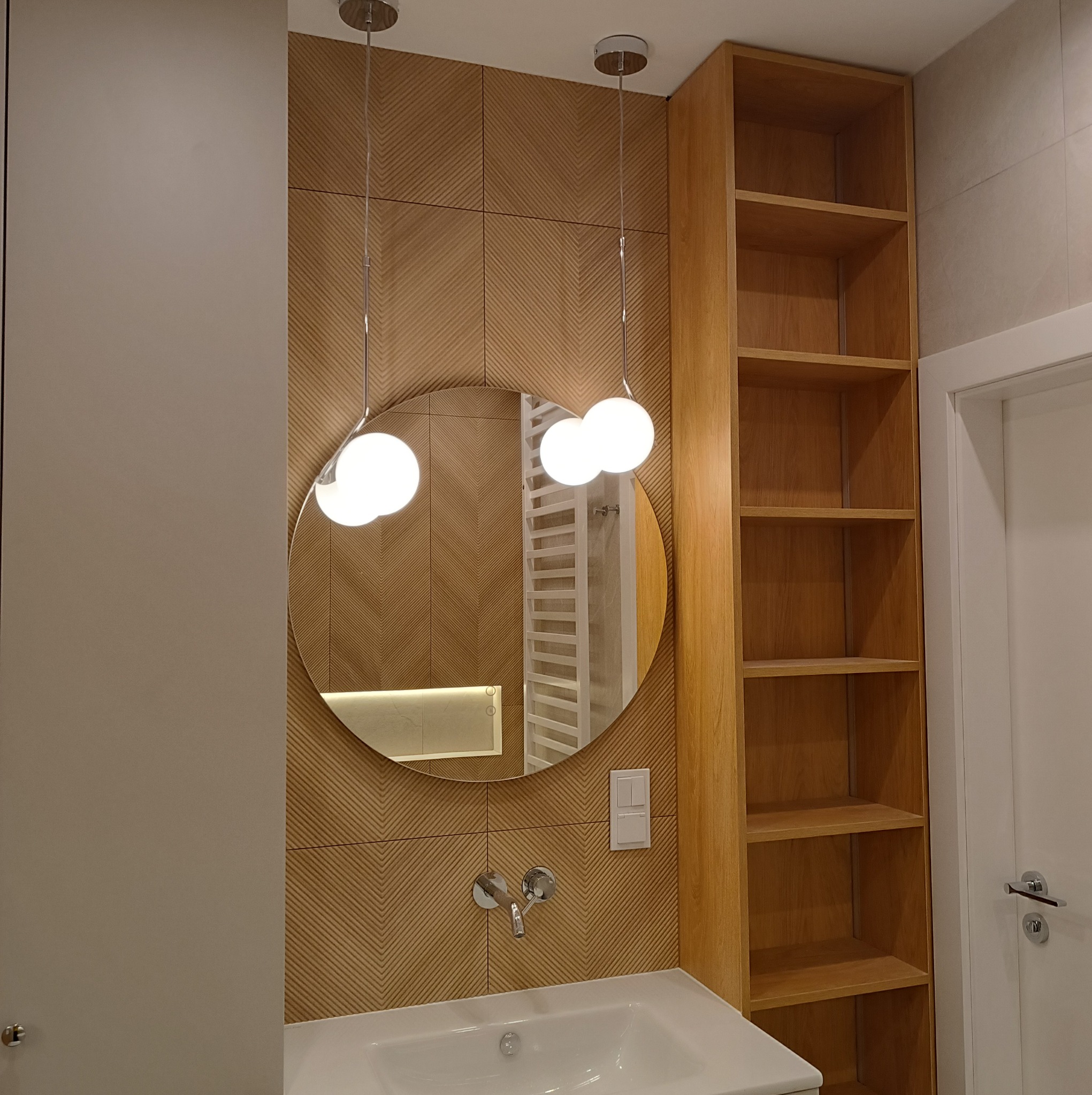 Nowoczesna łazienka z okrągłym lustrem, drewnopodobnymi płytkami w jodełkę, wiszącymi lampami kulami i drewnianym regałem z półkami.