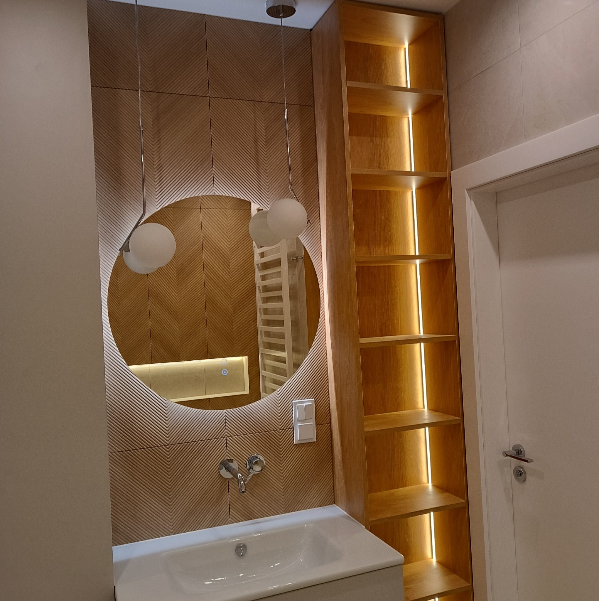 Nowoczesna łazienka z umywalką, okrągłym lustrem z podświetleniem, drewnianą szafką z oświetleniem LED i dekoracyjną ścianą z płytek ułożonych w jodełkę.