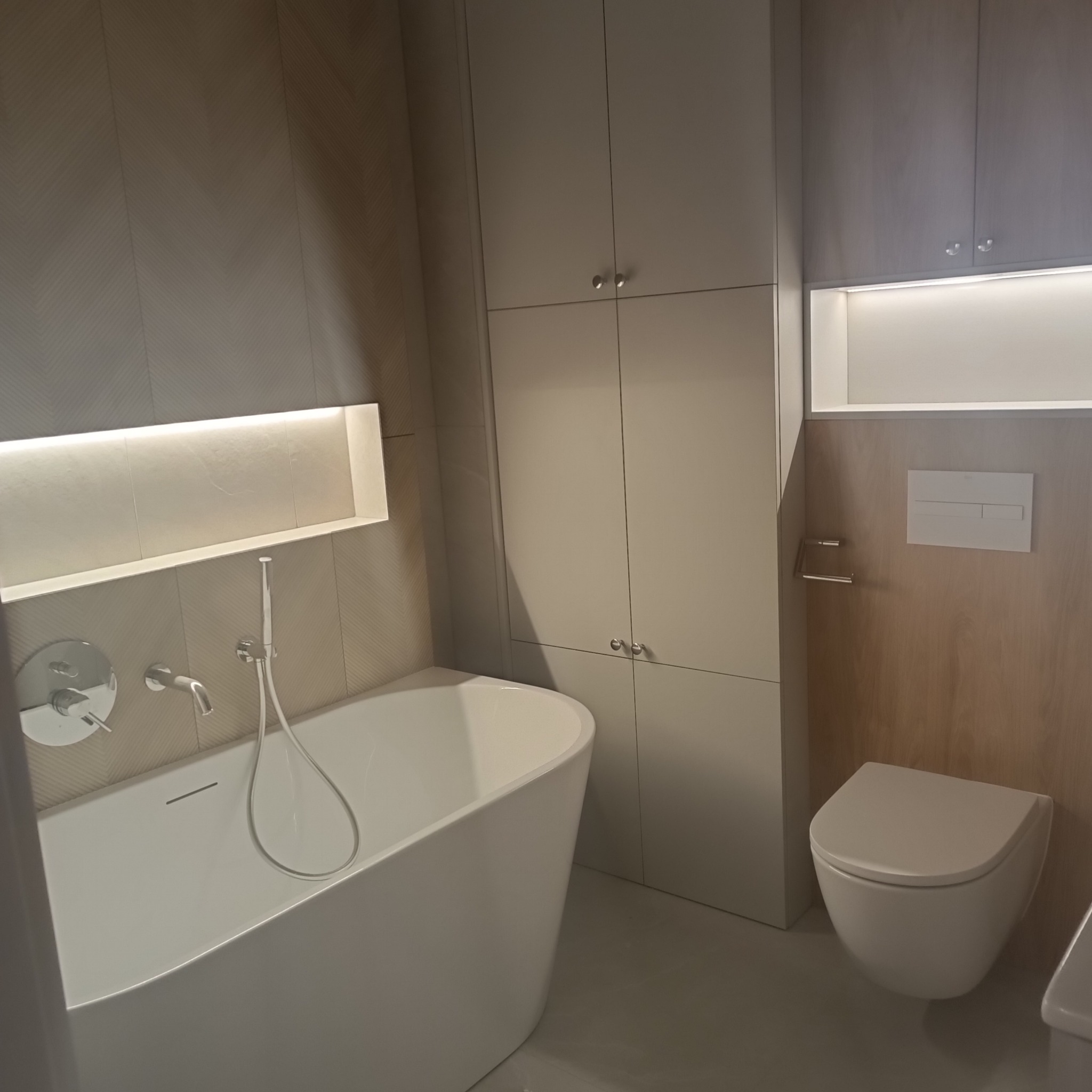 Nowoczesna łazienka z wolnostojącą wanną, zabudową meblową w odcieniach szarości i podświetlanymi niszami.