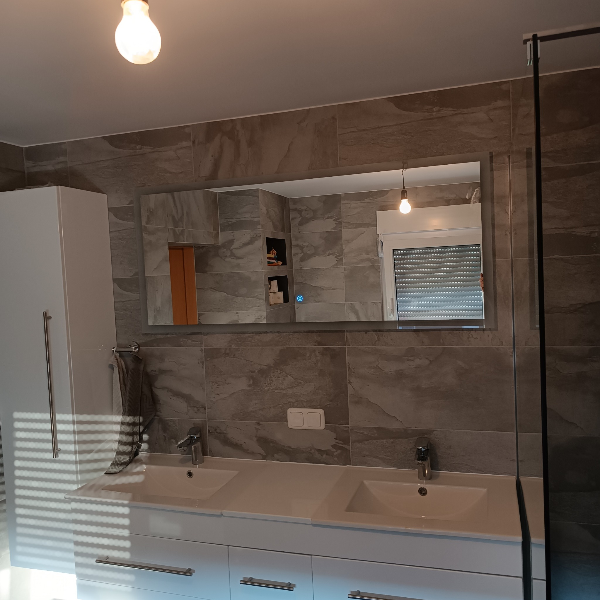 Nowoczesna łazienka w Nowogardzie z podwójną umywalką, dużym lustrem z podświetleniem LED i szarą płytką imitującą kamień. W lustrze odbija się okno z żaluzjami.