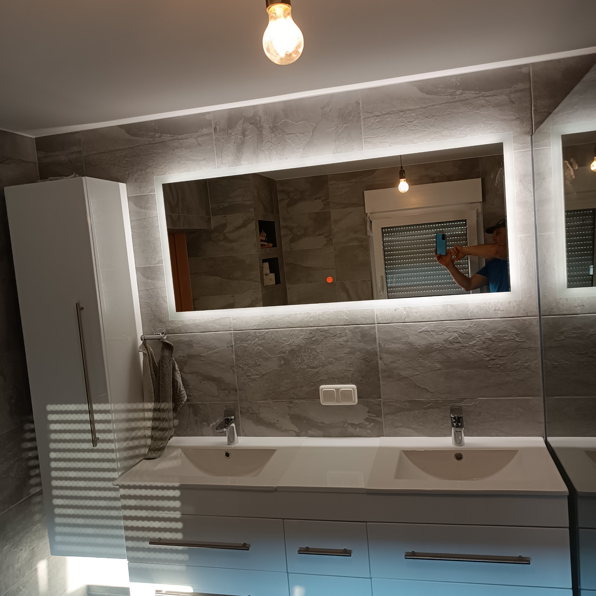 Nowoczesna łazienka z podwójną umywalką na białej szafce, szare płytki imitujące kamień, podświetlane lustro, widoczne odbicie osoby robiącej zdjęcie telefonem.