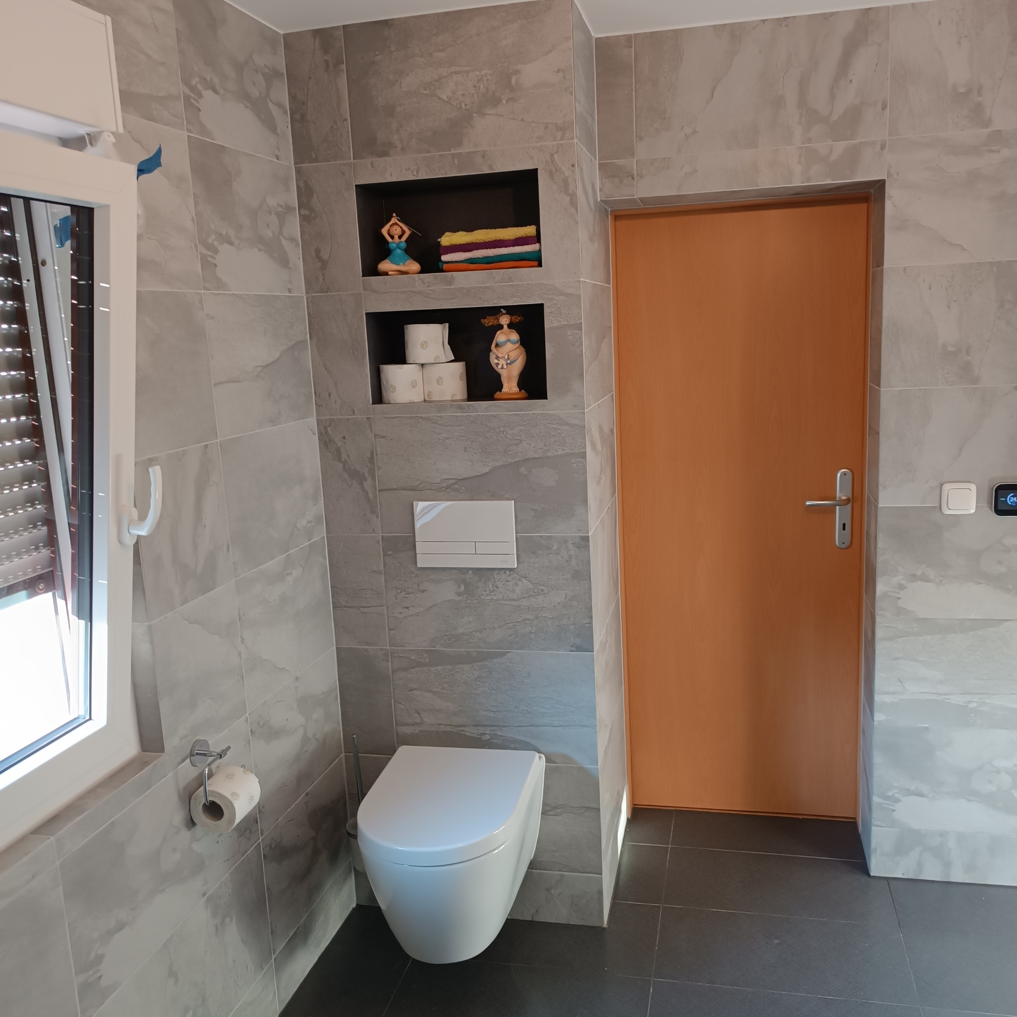 Nowoczesna łazienka z szarymi płytkami imitującymi marmur, podwieszana toaleta, drewniane drzwi i dekoracyjne wnęki z figurkami i ręcznikami.