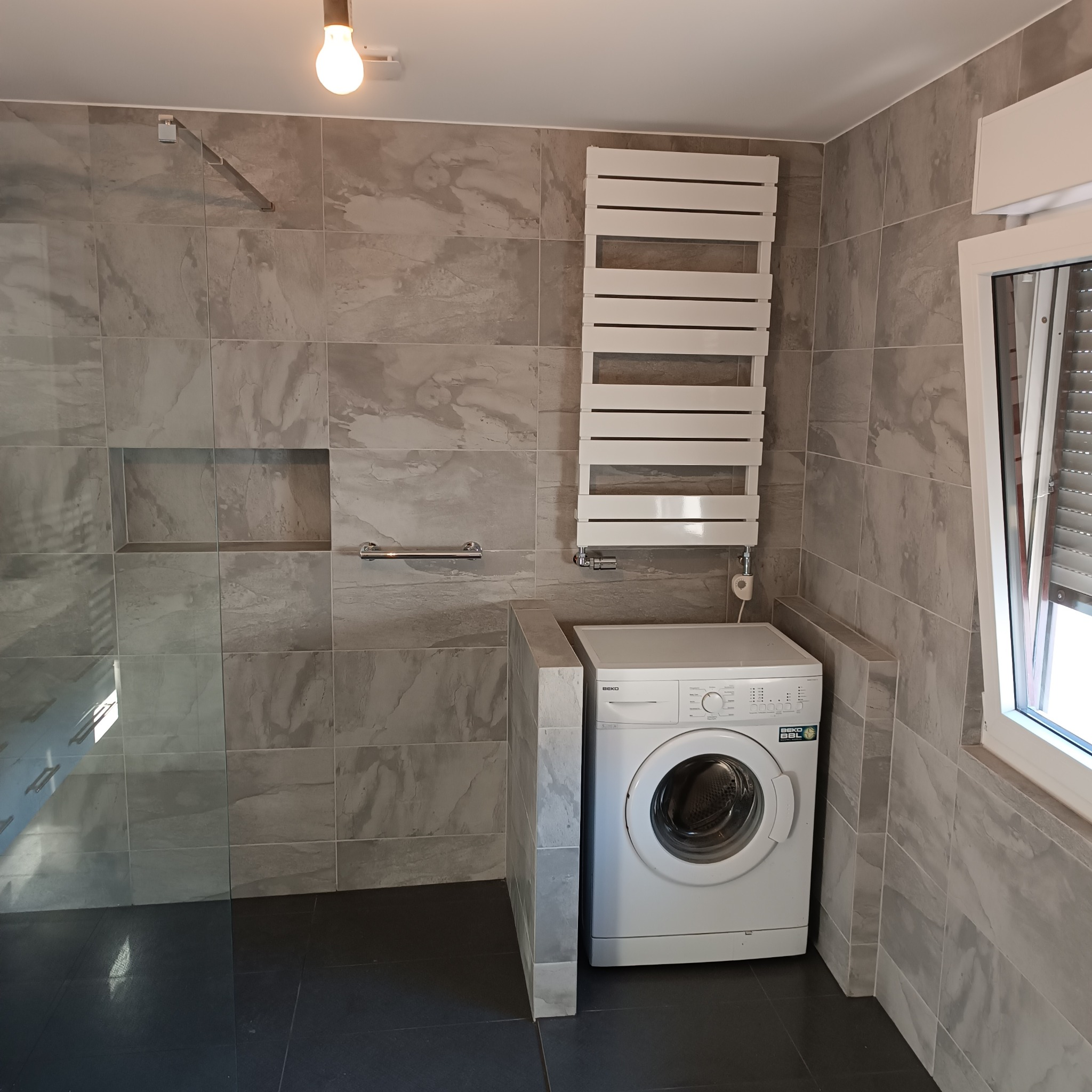 Szara łazienka z pralką, grzejnikiem drabinkowym i kabiną prysznicową typu walk-in, wykończona płytkami imitującymi marmur.