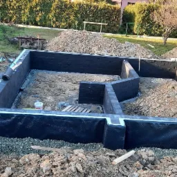 Fundamenty domu jednorodzinnego w Żywcu, zabezpieczone hydroizolacją, częściowo zasypane ziemią, widoczne bloczki fundamentowe i warstwa drenażowa.