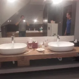 Nowoczesna łazienka z dwiema owalnymi umywalkami nablatowymi na drewnianym blacie, duże lustro z odbiciem osoby robiącej zdjęcie, widoczne akcesoria łazienkowe i dekoracyjne koszyki pod blatem.