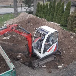 Kompaktowa koparka Neuson 2503 na gąsienicach, operator w kabinie, podczas prac ziemnych na działce z widoczną stertą ziemi i przyczepą, w tle rząd tuj