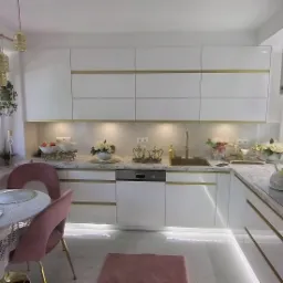 Elegancka, biała kuchnia z marmurowym blatem i złotymi akcentami, stół z różowymi krzesłami i dekoracjami kwiatowymi przy oknie.