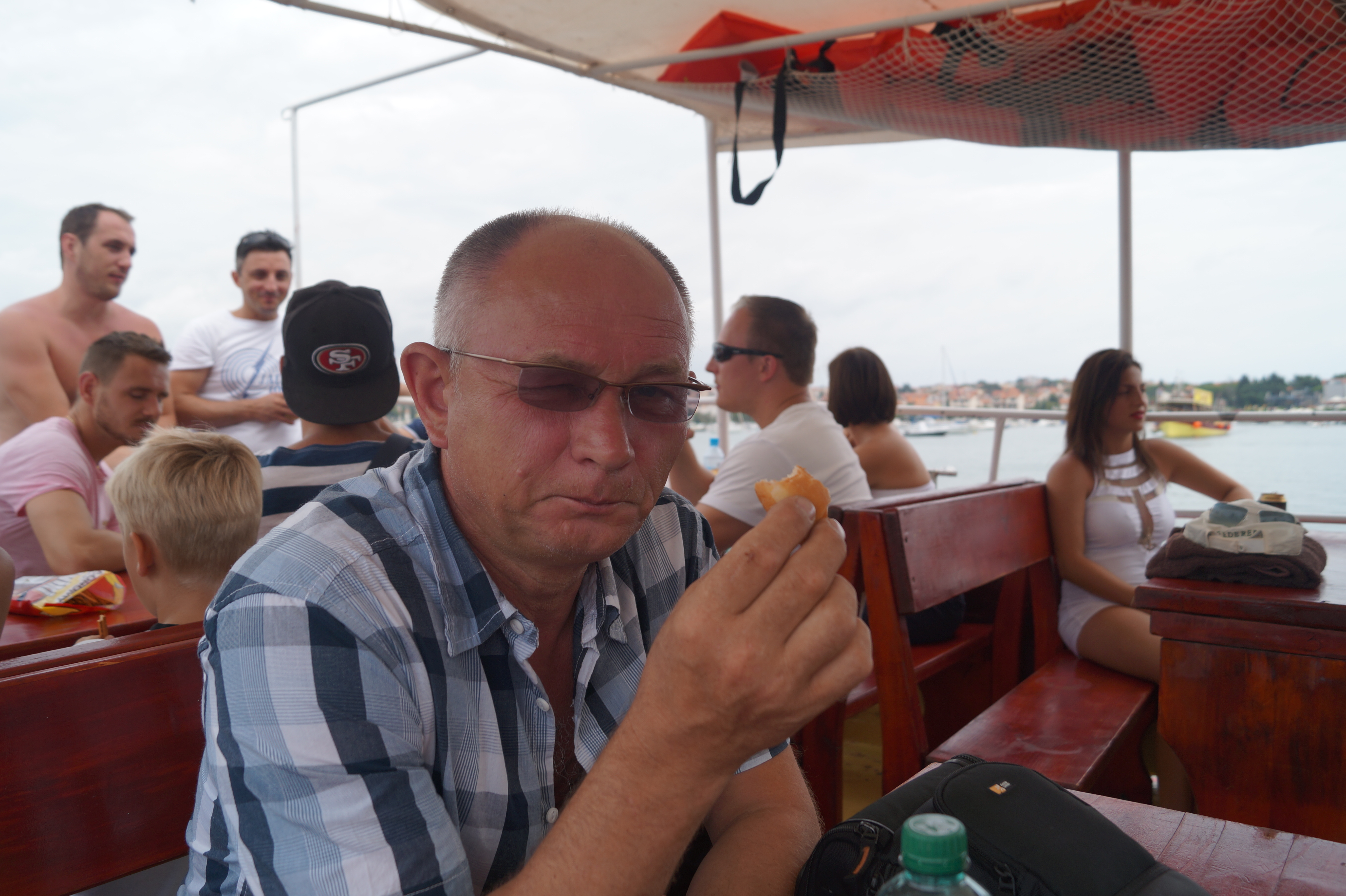 Mężczyzna w okularach i koszuli w kratę je pączka na łodzi, widok na morze i miasto w tle, inni pasażerowie siedzą przy stołach.