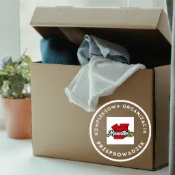 Otwarty karton z częściowo widoczną zawartością (ubrania, mata), obok doniczka z zieloną rośliną, na kartonie naklejka z logo firmy Manatki24 oferującej kompleksową organizację przeprowadzek.