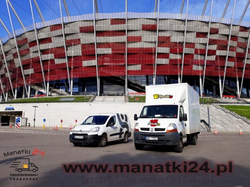 Dwa białe busy firmy Manatki24.pl zaparkowane na parkingu przed Stadionem Narodowym w Warszawie.