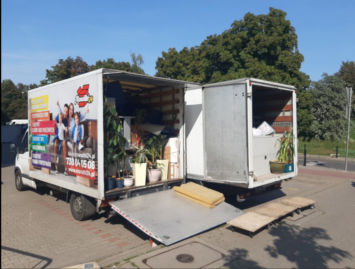 Ciężarówka z otwartą przestrzenią ładunkową, wypełnioną meblami i roślinami doniczkowymi, zaparkowana w Wołominie, z widocznym logo firmy Manatki24.pl na boku.