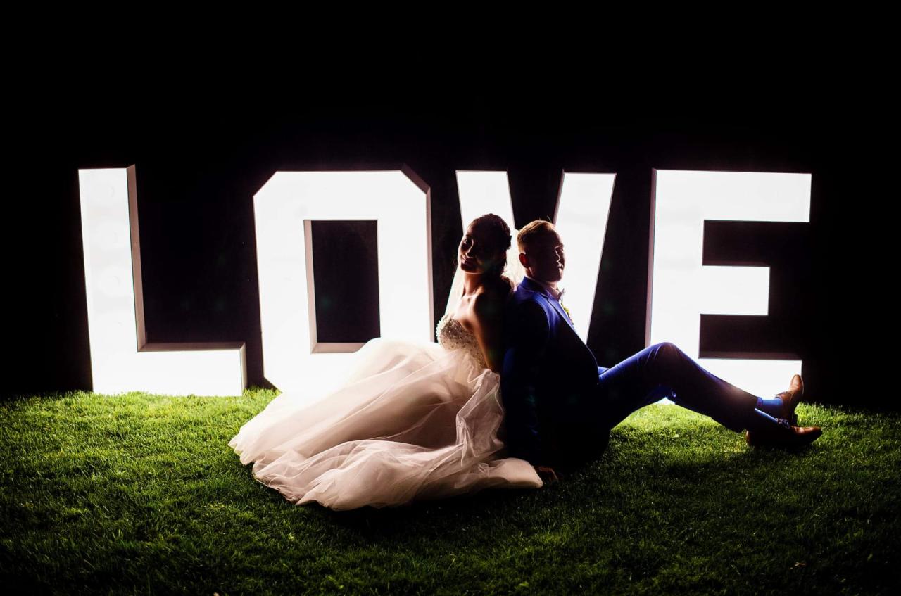 Para młoda siedząca na trawie oparta plecami o podświetlany napis LOVE, on w niebieskim garniturze, ona w białej sukni ślubnej z odkrytymi ramionami.