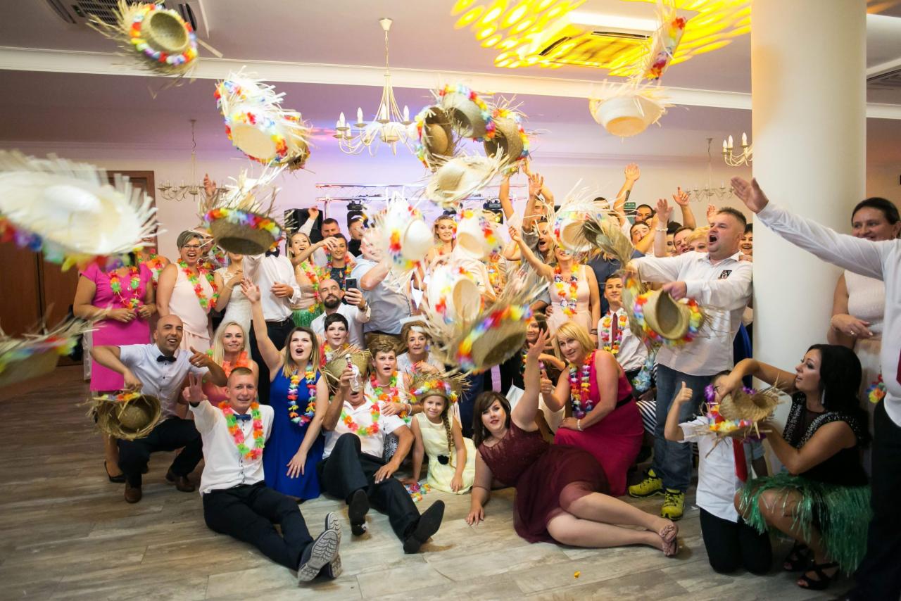 Grupa gości weselnych w hawajskich strojach rzuca w górę słomkowe kapelusze z kwiatami, świętując w sali z kolumną i żyrandolami.