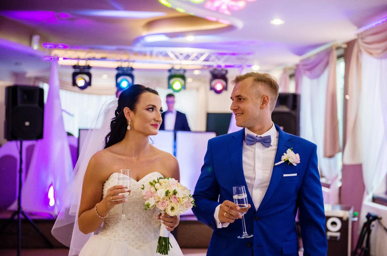 Para młoda w Cielczy, trzymająca kieliszki z szampanem, z DJ-em i sprzętem nagłośnieniowym w tle. Panna młoda w sukni bez ramiączek trzyma bukiet, a pan młody w niebieskim garniturze z muszką.