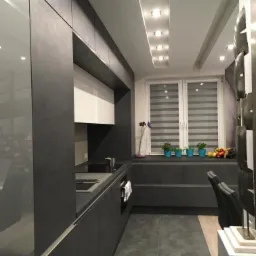 Nowoczesna kuchnia w odcieniach szarości z matowymi frontami, białymi szafkami wiszącymi i roślinami w niebieskich doniczkach na parapecie okiennym.