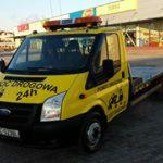 Żółta laweta Ford Transit z napisem 'Pomoc Drogowa 24H' zaparkowana na parkingu przed budynkiem sklepu. Widoczne światła ostrzegawcze na dachu.