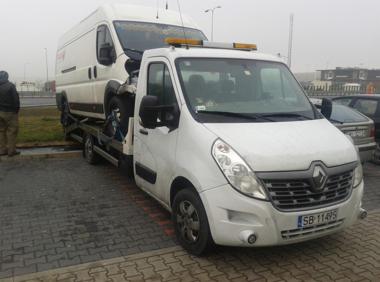 Biała laweta Renault z uszkodzonym białym busem na platformie, zaparkowana na brukowanym placu, w tle widoczny parking i budynek McDonald's w pochmurny dzień.