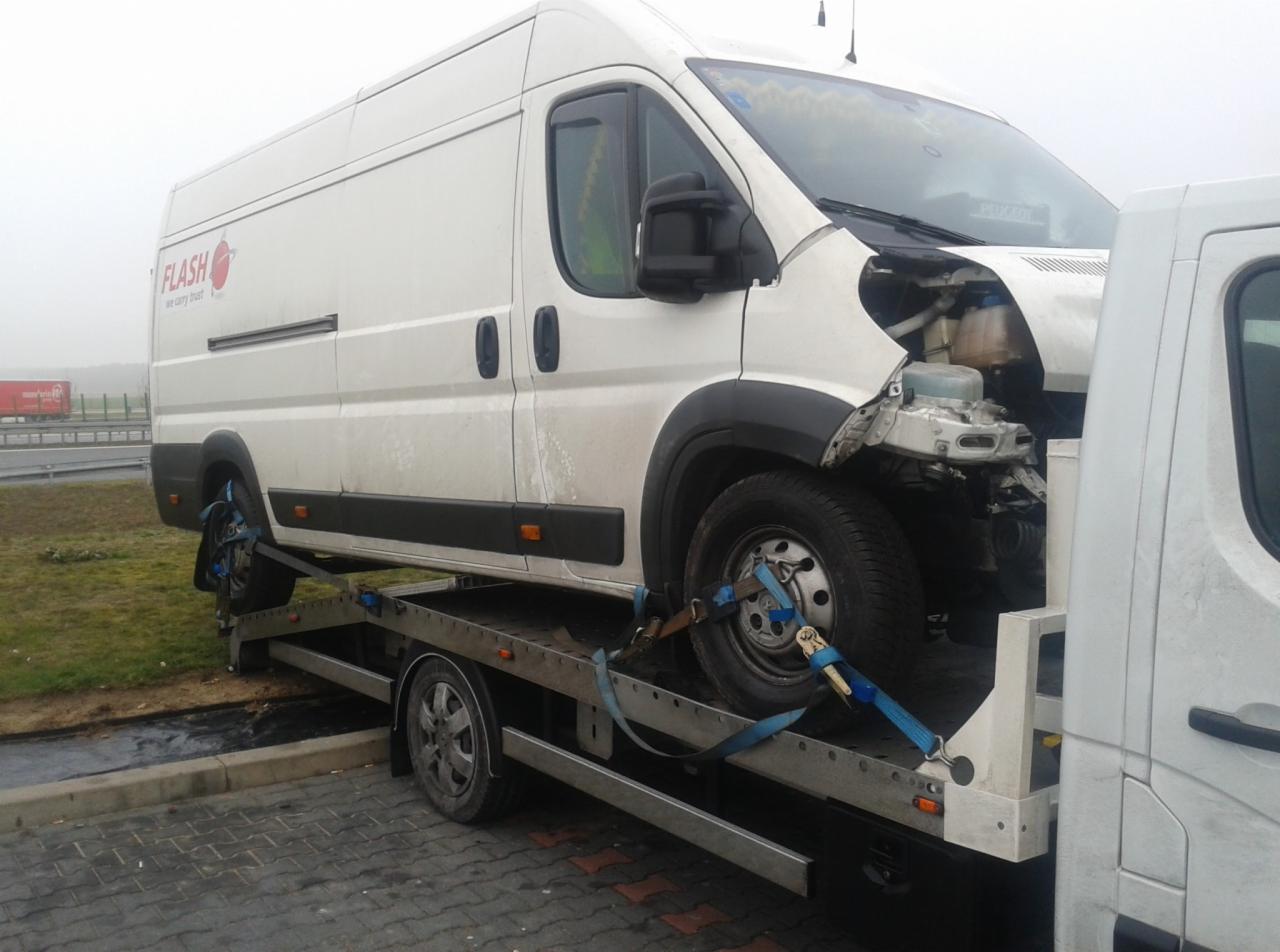 Uszkodzony biały bus marki Peugeot z logo Flash na lawecie, zabezpieczony niebieskimi pasami transportowymi, widoczny fragment autostrady w tle.