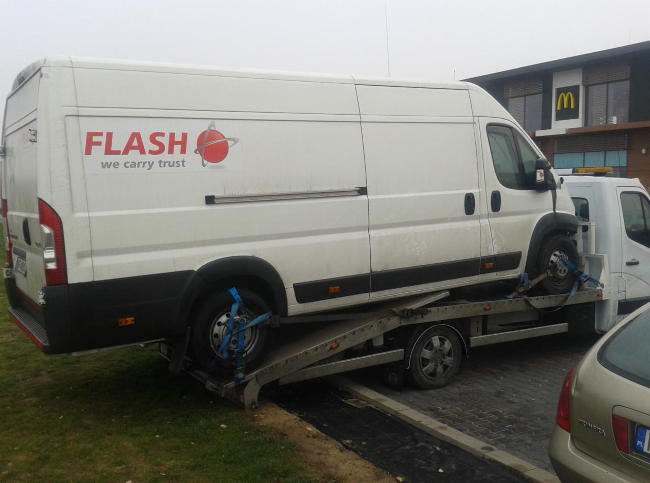 Biały dostawczy samochód marki Fiat z logo firmy transportowej FLASH umieszczony na platformie lawety, zabezpieczony niebieskimi pasami transportowymi, na tle budynku McDonald's.