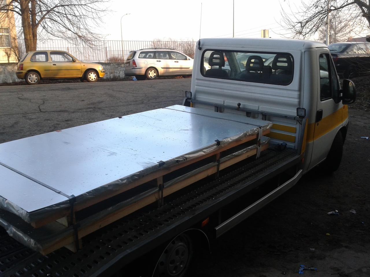 Laweta z ładunkiem blach zabezpieczonych drewnianymi belkami i folią, zaparkowana na tle miejskiego krajobrazu z żółtym i srebrnym samochodem osobowym.