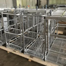 Gotowe, aluminiowe platformy robocze na kółkach, ułożone na palecie w hali produkcyjnej. Widoczne spawane konstrukcje z siatkową podłogą i pojemniki z płynami w tle.