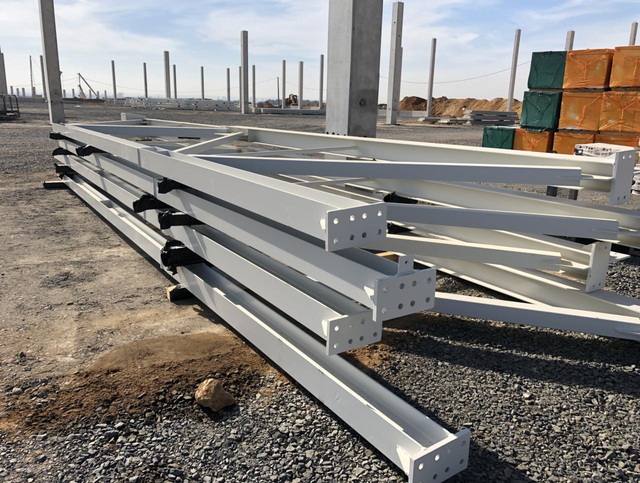 Stalowe, pomalowane na biało elementy konstrukcyjne hali, złożone na placu budowy, widoczne betonowe słupy w tle.