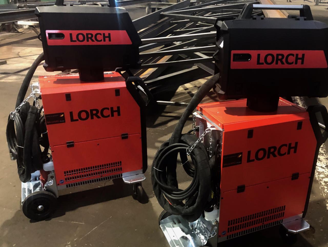 Dwa identyczne, pomarańczowo-czarne, mobilne urządzenia spawalnicze marki Lorch z przewodami, na tle konstrukcji stalowej.