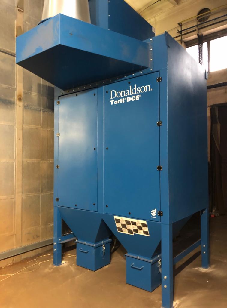 Stacjonarny, niebieski odpylacz przemysłowy Donaldson Torit DCE z metalową konstrukcją nośną i filtrem workowym, umieszczony w hali produkcyjnej.