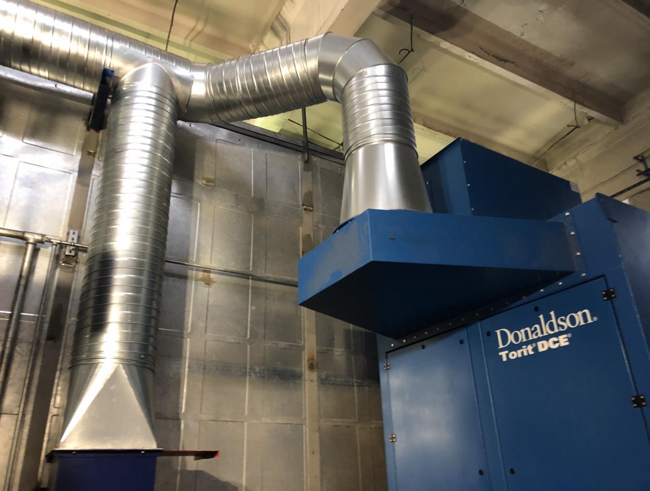 System odpylania Donaldson Torit DCE z widocznymi, metalowymi rurami wentylacyjnymi w hali przemysłowej.