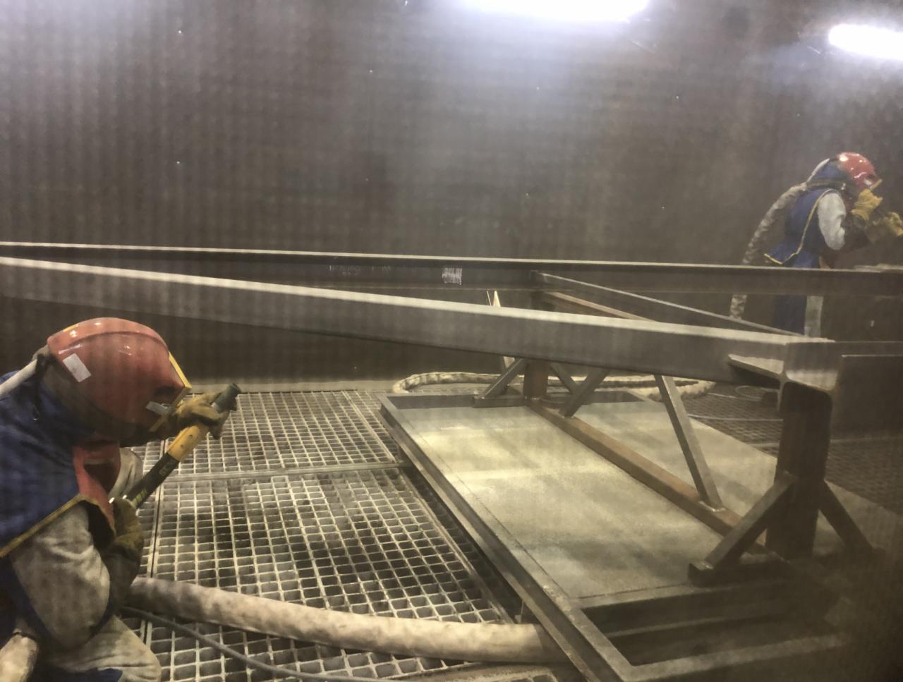 Pracownik w kombinezonie ochronnym i masce piaskuje stalową konstrukcję wewnątrz kabiny, widoczne iskry i pył.