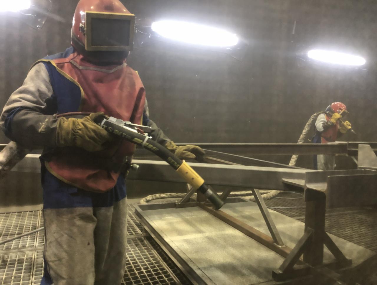 Pracownik w kombinezonie ochronnym i masce piaskuje metalową konstrukcję w hali przemysłowej, drugi pracownik w tle wykonuje te same czynności.