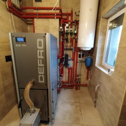 Ajbm - Kotłownia z piecem Defro ES 10, czerwonymi rurami instalacji grzewczej, bojlerem i oknem, widok na całą instalację.