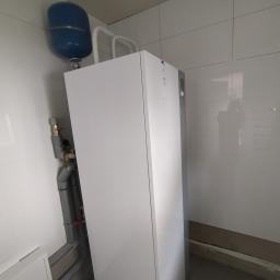 Ajbm - Nowoczesny, biały piec gazowy z niebieskim zbiornikiem wyrównawczym, zamontowany na tle białych płytek ceramicznych, widoczne rury instalacji.