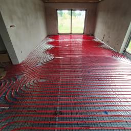 Ajbm - Czerwone rury ogrzewania podłogowego ułożone na czarnej siatce w surowym pomieszczeniu z widokiem na ogród przez okno.