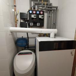 Ajbm - Kompaktowa kotłownia: biały piec Viessmann, szary system rur z izolacją, czarny panel kontrolny z manometrami, niebieski zbiornik wyrównawczy, oraz zmiękczacz wody w pomieszczeniu wyłożonym białymi...