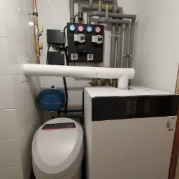 Kompaktowa kotłownia: biały piec Viessmann, szary system rur z izolacją, czarny panel kontrolny z manometrami, niebieski zbiornik wyrównawczy, oraz zmiękczacz wody w pomieszczeniu wyłożonym białymi...