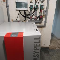Ajbm - Nowoczesny piec na pellet EasyPell z widocznymi rurami instalacji grzewczej, pompami obiegowymi Wilo i manometrem w kotłowni o surowym wykończeniu.